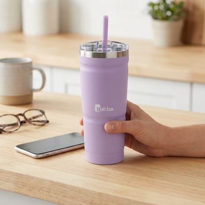 A hand holds the bubba Envy S Stainless Steel Tumbler in Lavender Frost on a kitchen counter, providing a clear scale reference next to a smartphone and other common items.