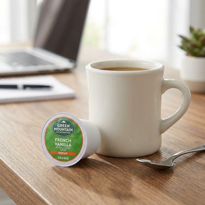 A Keurig Green Mountain French Vanilla Decaf K-Cup pod rests on a wooden desk next to a standard ceramic coffee mug for size reference.