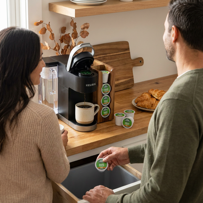A couple in a kitchen uses a Keurig machine, with Green Mountain French Vanilla Decaf pods visible in a holder and one being placed in a waste bin.
