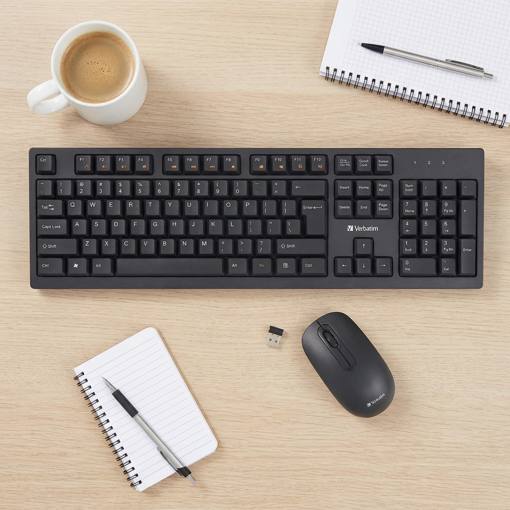 The Verbatim Wireless Keyboard and Mouse on a wooden desk, showing scale next to a coffee mug and notepads.