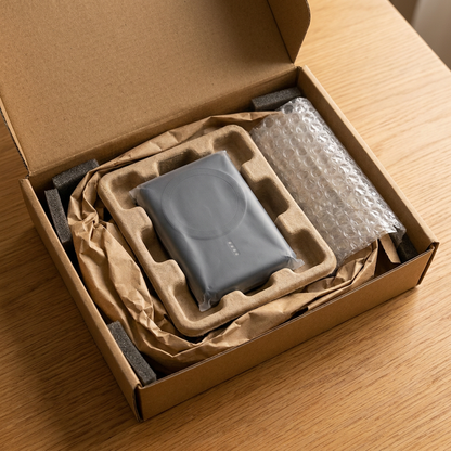 A photograph shows the Verbatim magnetic power bank from the reference image inside an open cardboard shipping box. The device is securely nestled in a fitted molded pulp tray and a plastic sleeve, surrounded by crushed paper and a bubble wrap roll, all on a wooden surface.