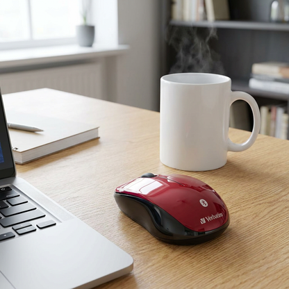 A photograph showing the red Verbatim Bluetooth mouse on a wooden desk next to a white ceramic coffee mug to demonstrate its compact size.
