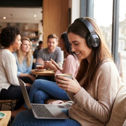 A woman in a beige sweater smiles with Sharp HP-60 headphones in a busy cafe, looking at a laptop.
