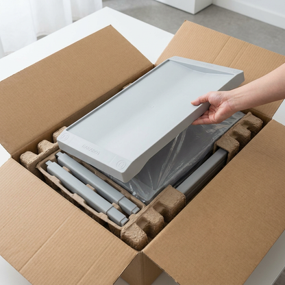 A person is unboxing the Rakabot Plastico 2-tier shoe rack, lifting one grey plastic shelf from a cardboard box. The parts are securely nestled within molded pulp packing and protective wrap.