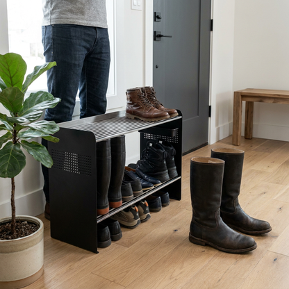 A polished product photograph shows the RAKABOT Signature 2-tier shoe rack in an entryway, made of matte black metal and stainless steel. An adult person in jeans stands next to it, providing scale. The rack holds various shoes, including tall boots, with more boots on the wooden floor beside it.